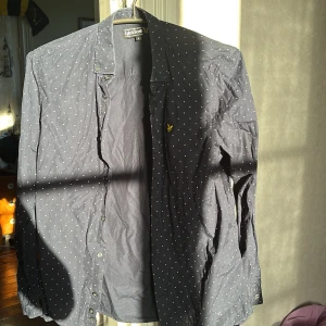 Mörkblå prickig skjorta Lyle & Scott - Snygg mörkblå skjorta från Lyle & Scott med vita prickar och klassisk krage. Skjortan har långa ärmar, knäppning framtill och den ikoniska gula loggan broderad på bröstet. Perfekt för dig som gillar stilrena detaljer och mönster.