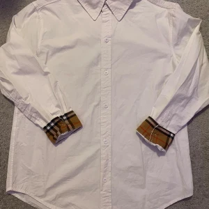 Vit skjorta b - Vit skjorta med klassisk krage och knäppning framtill. Manschetterna har den ikoniska rutiga insidan i beige, svart och rött. Skjortan är långärmad och i bomull, perfekt för en clean och stilren look. Storlek L men sitter som en M