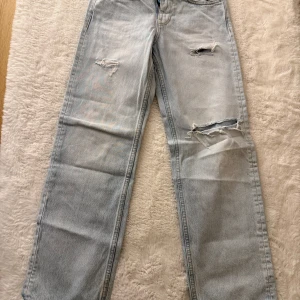 Ljusblå raka jeans med slitningar - Säljer ett par ljusblå jeans med raka ben och snygga slitningar på låren och knät. Jeansen har klassisk femficksdesign och är tillverkade i bomullsdenim. Perfekta för en avslappnad och trendig look.