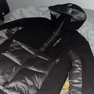 Svart dunjacka Sail Racing med huva - Säljer en ny svart dunjacka från Sail Racing med glansiga paneler och stor reflekterande logga på ryggen. Jackan har huva, dragkedja framtill och är fylld med 600 FP dun. Perfekt för kalla dagar