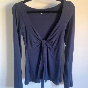 Mörkblå långärmad topp med knyt - Snygg mörkblå långärmad topp med djup v-ringning och knyt framtill. Toppens material är mjukt och stretchigt, vilket gör den bekväm att bära. Perfekt för dig som gillar en enkel men trendig look.