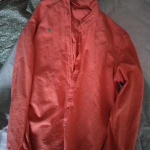 Korallröd henley-skjorta med lång ärm - Säljer en korallröd henley-skjorta med lång ärm och ståkrage. Skjortan har en liten grön broderad detalj på bröstet och är tillverkad i ett mjukt bomullstyg. Perfekt för dig som gillar en avslappnad men ändå stilren look.