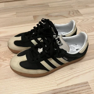 Adidas Samba OG svart/vit sneakers - Säljer ett par klassiska Adidas Samba OG sneakers i svart och vitt med beige detaljer. Skorna har ikoniska tre ränder på sidan, svart ovandel i skinn och mocka, samt gummisula. Perfekta för dig som gillar retrostil och streetwear. Skön passform och snygg design.