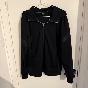 Svart hoodie från BOSS med dragkedja - Svart hoodie från BOSS med hel dragkedja, huva och diskreta logodetaljer på bröstet och ärmarna. Tröjan har långa ärmar, snörning i huvan och ribbade muddar. Perfekt för en chill och stilren look.