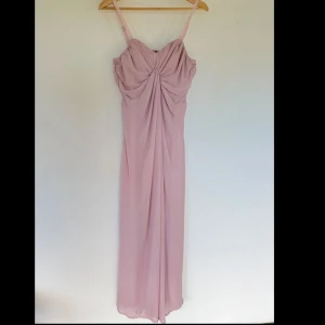 Ljusrosa långklänning med drapering - Elegant ljusrosa långklänning med smala axelband och snygg drapering framtill. Klänningen har en feminin siluett och ett mjukt, flytande tyg som ger en romantisk känsla. Perfekt för speciella tillfällen när du vill känna dig extra fin. Står att storleken är 46 men är väldigt liten i storlek skulle säga M/L. Måtten/ byst 94cm, midja 86cm och längden på klänningen är 125 cm. Klänningen är även lite stretchig.