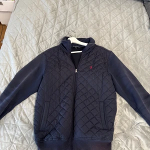 Mörkblå quiltad jacka Polo Ralph Lauren - Snygg mörkblå quiltad jacka från Polo Ralph Lauren med dragkedja framtill och ribbade muddar. Jackan har långärmade ärmar, två fickor och den klassiska röda loggan broderad på bröstet. Perfekt för dig som gillar stilren och sportig look.