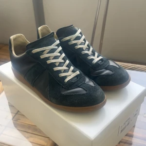 Maison Margiela blåa sneakers i mocka - Maison Margiela sneakers i blåt mocka och skinn med vita snören och gummisula. Klassisk low-top siluett med rund tå och kontrasterande detaljer. Kommer med originalkartong och dustbags. Perfekta för dig som gillar stilrena och lyxiga sneakers.