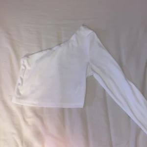 Vit ribbad one shoulder topp - Säljer en vit ribbad topp med one shoulder-design och lång ärm. Toppen är croppad och har en tight passform som framhäver siluetten. Perfekt för dig som gillar trendiga och stilrena plagg med en twist.