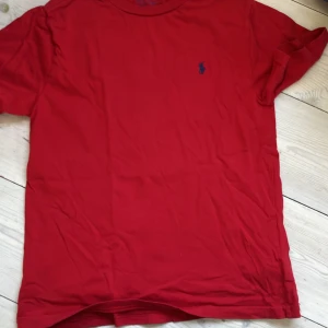 Röd t-shirt från Ralph Lauren, strl 10-12 - Röd t-shirt från Ralph Lauren med klassisk passform och blå broderad logga på bröstet. Tillverkad i mjuk bomull och har korta ärmar samt rund halsringning. Perfekt för en stilren och enkel look.