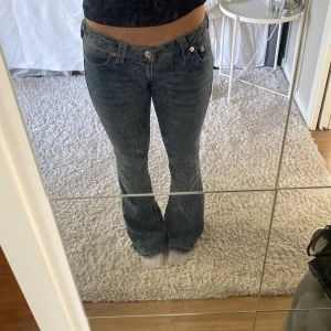 True Religion bootcut jeans blå - Snygga blå jeans från True Religion med klassisk bootcut-modell och låg midja. Jeansen har fem fickor, silverfärgade nitar och detaljerade sömmar. Tillverkade i mjukt denim med lätt tvättad look och logga bak på linningen.