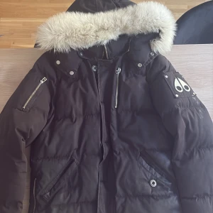 Svart Moose Knuckles dunjacka med päls - Säljer en svart dunjacka från Moose Knuckles med fluffig ljus päls runt huvan. Jackan har flera dragkedjor, logga på ärmen och broderad insida. Perfekt för kalla dagar och riktigt snygg streetstil.