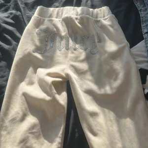 Beiga Juicy Couture mjukisbyxor med strass - Beiga mjukisbyxor från Juicy Couture i mjuk velour med resår och snörning i midjan. Baksidan pryds av ordet 'Juicy' i glittrande strass. Klassisk avslappnad passform och diskret logga framtill. Perfekta för en chill och trendig look.