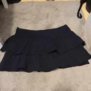 Mörkblå lågmidjad volangkjol. - Supersöt mörkblå kortkjol med två lager volanger och sitter lågt vid midjan. Kjolen har en flowy känsla och passar perfekt till sneakers eller boots. Enkel att matcha och ger en trendig look.