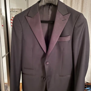 Svart kavaj med satinrevers - Stilren svart kavaj med breda satinrevers och två knappar framtill. Klassisk design med tre fickor och slät yta, perfekt för dig som vill ha en elegant look. Passar dig som gillar tidlös stil och vill sticka ut med snygga detaljer. 