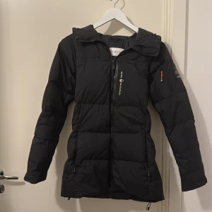 Sail Racing dunjacka svart - Svart dunjacka från Sail Racing. Jackan har huva, dragkedja framtill och flera praktiska fickor. Lång modell med quiltad design, perfekt för kalla dagar. Använd en vinter och har inga fläckar eller skador. Håller värme riktigt bra. Är storlek small men passar även som xs och m