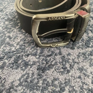 Svart skärp från Armani i skinn - Svart bälte från Armani i äkta skinn med stilren, metallfärgad spänne där loggan och märket är ingraverade. Bältet har en klassisk, rak form och passar perfekt till jeans eller kostymbyxor. Snygg detalj med diskret logotyp på spännet.