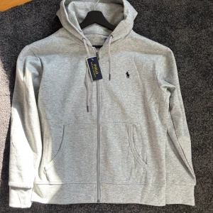 Grå Polo Ralph Lauren - Snygg ljusgrå hoodie från Polo Ralph Lauren med dragkedja, huva med snörning och klassisk broderad logga på bröstet. Tröjan har känguruficka och är tillverkad i mjuk bomullsmix. Perfekt för chill dagar eller när du vill ha en clean look.