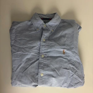 Blå skjorta från Polo Ralph Lauren - Klassisk ljusblå skjorta från Polo Ralph Lauren med broderad logga på bröstet. Skjortan har knappar framtill, button-down krage och långa ärmar. Tillverkad i mjuk bomull och passar perfekt till en clean och stilren look.