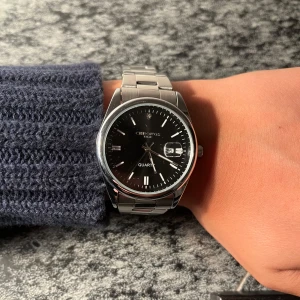 Svart Chronos klocka - Stilren klocka från Chronos med svart urtavla och datumvisning. Armbandet är i silverfärgat metall och har en klassisk länkdesign. Urtavlan är rund med tydliga indexmarkeringar och en diskret logga upptill. Perfekt accessoar för dig som gillar en clean look.