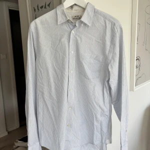 Randig skjorta från Acne Studios - Klassisk skjorta från Acne Studios i vitt med tunna blå ränder. Skjortan har lång ärm, bröstficka och knäppning framtill. Tillverkad i bomull för en skön och luftig känsla. Liten fläck på vänster bröstficka. Se bild. Hör av er om ni har några frågor 