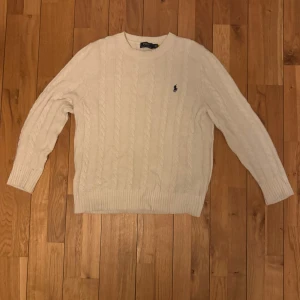 Vit kabelstickad tröja Polo Ralph Lauren - Kabelstickad vit tröja från Polo Ralph Lauren med klassisk rund halsringning och broderad marinblå logga på bröstet. Tröjan har lång ärm och ribbade muddar för en snygg passform. Perfekt för lager-på-lager under kyliga dagar.