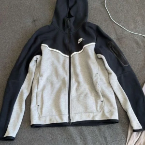 Nike tech fleece tvåfärgad - Nike hoodiejacka i svart och ljusgrått med hel dragkedja och huva. Jackan har en vit Nike-logga på bröstet, praktiska fickor och en extra ficka med dragkedja på ärmen. Sportig design med kontrastpaneler och bekvämt material.