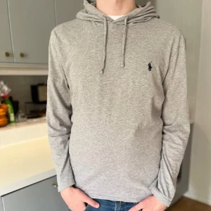 Grå hoodie från Ralph Lauren - Snygg grå hoodie från Ralph Lauren med klassisk huva och snörning. Tröjan har lång ärm och en liten mörkblå logga broderad på bröstet. Tillverkad i mjuk bomull 