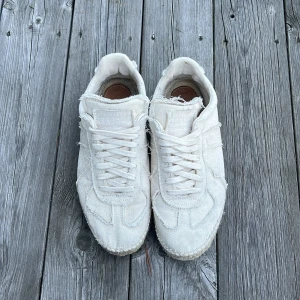 Maison margiela espadrilles - Säljer ett par vita sneakers med fransiga kanter och snörning. Skorna har en unik espadrille-inspirerad sula med grov söm runt om. Tillverkade i bomullstyg med platt sula och rund tå. Perfekta för dig som gillar en chill och avslappnad stil.
