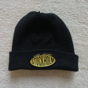 Svart mössa Marc Jacobs Stockholm - Svart ribbstickad mössa från Marc Jacobs med gul och svart broderad patch framtill där det står 'STOCKHOLM'. Klassisk form med uppvikt kant, perfekt för att hålla värmen och ge en cool streetstyle-vibe. Har ett litet hål upptill. 