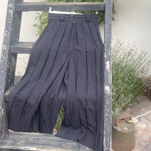 Svarta vida kostymbyxor Esther Jane - Säljer ett par svarta kostymbyxor från Esther Jane London. Byxorna är högmidjade med vida ben och snygga pressveck. De har bälteshällor, två knappar och dragkedja framtill. Perfekta för dig som gillar en loose och trendig siluett.