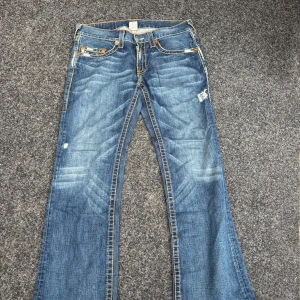 True Religion blå bootcut jeans - Säljer ett par blå jeans från True Religion med bootcut passform. Jeansen har slitna detaljer, kontrastsömmar och ikoniska broderade bakfickor med orange och gul tråd. Klassisk femficksmodell i denim med dragkedja och knapp. Perfekta för dig som gillar streetstyle.
