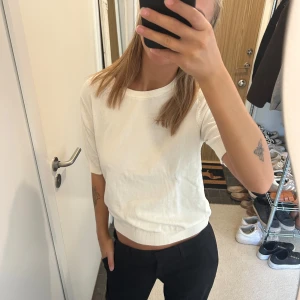 Vit kortärmad topp i bomull - Säljer en enkel och stilren vit topp med korta ärmar och rund hals. Toppen är något croppad och har en mjuk, ribbad kant nedtill och vid ärmsluten. Perfekt för dig som gillar en clean och klassisk look.