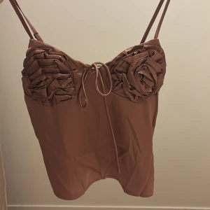 Brun satinlinnetopp med rosdetaljer - Supersöt brun linnetopp i satin med smala axelband och unika rosdetaljer på bysten. Toppen har en snygg knytning framtill och en glansig finish som ger en lyxig känsla. Perfekt för dig som vill sticka ut med en trendig och romantisk look.