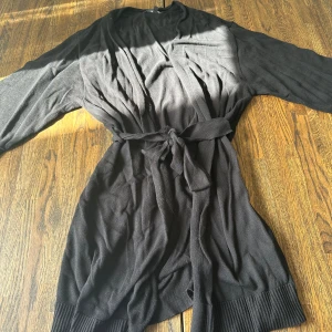 Svart kimono från Happy Holly - Svart kimono från Happy Holly med knytband i midjan och långa ärmar. Cardiganen är stickad och har breda ribbade muddar vid ärmslut och nederkant. Perfekt för dig som gillar en enkel men snygg look.