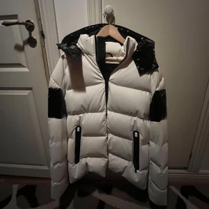 Vit och svart pufferjacka Moose Knuckles - Snygg pufferjacka från Moose Knuckles i vit och svart med glansiga svarta detaljer på huva, ärmar och rygg. Jackan har dragkedja framtill, två fickor med svarta detaljer och stor huva. Jackan kom aldrig till användning därför säljs. Jackan är aldrig använd.