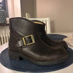 Johnny Bulls bruna boots i skinn - Säljer ett par klassiska bruna Johnny Bulls boots i äkta skinn med robust sula och snyggt spänne i mässing. Modellen har rund tå och låg klack, perfekt för dig som gillar en tidlös och cool look. Märket är präglat på sidan och sulan. Sparsamt använda 