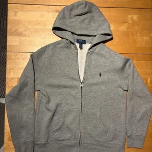 Grå hoodie från Polo Ralph Lauren - Grå hoodie från Polo Ralph Lauren med dragkedja och klassisk broderad logga på bröstet. Tröjan har huva, känguruficka och är tillverkad i mjuk bomullsblandning. Perfekt för chill dagar eller när du vill ha en clean och enkel look.