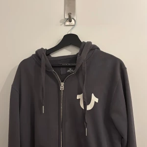 Mörkgrå hoodie från True Religion - Snygg mörkgrå hoodie från True Religion med dragkedja och vit logga på bröstet. Tröjan har huva med snörning och är tillverkad i mjuk bomull. Perfekt för en avslappnad och cool stil.