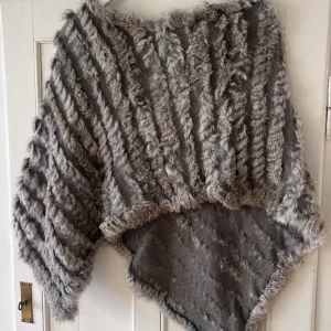 Grå pälsstickad ponchotröja - Supermysig grå ponchotröja med pälsdetaljer och stickad struktur. Tröjan har bred passform och är perfekt för kyliga dagar. Snygga diagonala ränder och fluffig känsla gör den till ett statement-plagg.