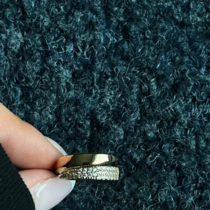 Guldig ring med glittrande stenar - Snygg guldig ring med en bred, slät yta och en rad glittrande stenar framtill. Ringen har en modern och stilren design som verkligen drar blickarna till sig. Perfekt för dig som gillar att sticka ut med dina accessoarer.
