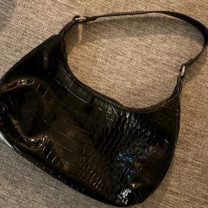 Svart axelväska i croco-imitation - Snygg svart axelväska med glansig croco-imitation och stilren form. Perfekt storlek för det viktigaste och enkel att slänga över axeln. Väskan har ett trendigt och tidlöst utseende som passar till många outfits.