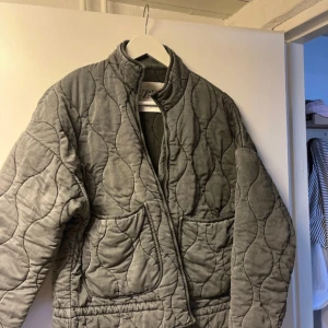 Grå quiltad jacka från Zara XS - Quiltad grå jacka från Zara i storlek XS. Jackan har en loose passform, ståkrage och stora fickor framtill. Snygg vågig quiltning och mjukt material som känns skönt mot huden. Perfekt för lager-på-lager under höst och vår.