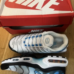 Nike Air Max Plus blå/vit sneakers - Nike Air Max Plus sneakers i vitt och blått med svarta detaljer och ikoniska vågformade linjer. Skorna har synlig Air Max-dämpning, mesh-överdel och svarta yttersulor. Swoosh-logga på sidan och blå detaljer vid snörningen. Perfekta för dig som gillar streetwear och snygg design.