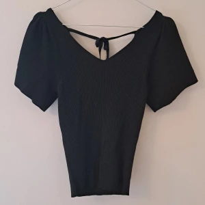 Svart ribbad topp med knyt i ryggen - Svart ribbad topp med korta puffärmar och v-ringning. Toppen har en snygg knytning i ryggen och är figurnära i modellen. Perfekt för dig som gillar stilrena och trendiga plagg med det lilla extra.