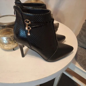 Svarta stövletter med ormmönster - Snygga svarta stövletter med spetsig tå och hög klack. Skorna har ett ormmönstrat material och guldfärgade detaljer på sidan. Dragkedja baktill för enkel påtagning. Perfekta för dig som vill sticka ut med en edgy look.