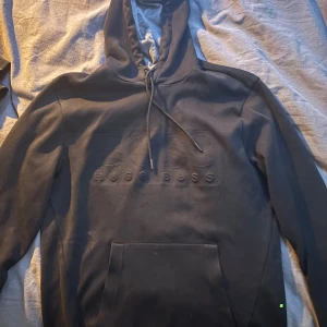 Svart hoodie från Hugo Boss - Svart hoodie från Hugo Boss med broderad logga framtill. Klassisk huva med snörning och stor magficka. Mjuk bomullsblandning och långärmad modell, perfekt för en avslappnad och stilren look.