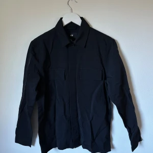 Svart overshirt från H&M, strl 46 - Svart overshirt från H&M i storlek 46. Skjortan har klassisk krage, två bröstfickor med lock och knäppning framtill. Materialet är 70% bomull och 30% polyamid, vilket ger en skön och lite robust känsla. Perfekt att slänga över en t-shirt för en chill look. 3S19
