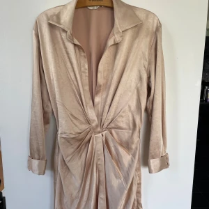 Champagnefärgad festklänning från abyss by abby the label  - Snygg champagnefärgad kort festklänning från abyss by abby the label. Storlek xs, snygg åtsittande passform med lång ärm, markerad midja och coola knappar vid ärmsluten. Klänningen har omlottkänsla framtill och är gjord i ett glansigt, mjukt material som ger en lyxig vibe. Använd 1 gång så i nyskick🥰