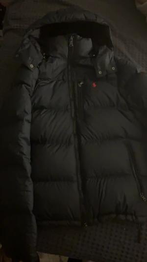Svart dunjacka från Polo Ralph Lauren Äkta - Svart dunjacka från Polo Ralph Lauren med röd broderad logga på bröstet. Jackan har huva, flera dragkedjor och en puffig siluett som håller dig varm. Perfekt för kalla dagar och har en clean, stilren look.