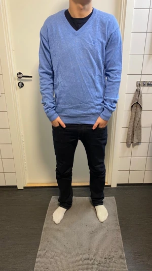 Blå v-ringad tröja från Gant - Snygg ljusblå v-ringad tröja från Gant med diskret logga på bröstet. Tröjan är i mjuk bomull, har långa ärmar och ribbade muddar vid ärmslut och nederkant. Perfekt för lager-på-lager och passar till jeans eller chinos.